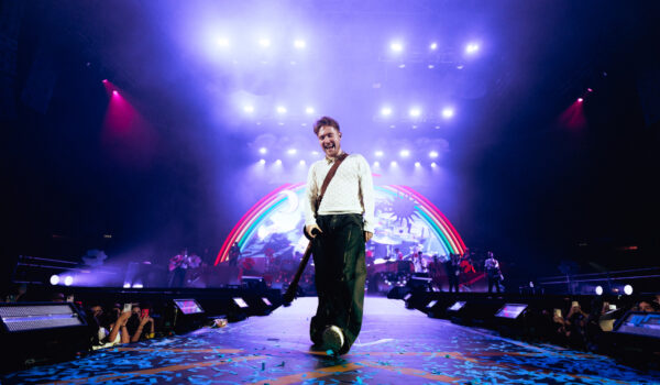 il cantante Alfa su pedana davanti al palco durante un concerto; luci sul soffito e arcobaleno sullo sfondo