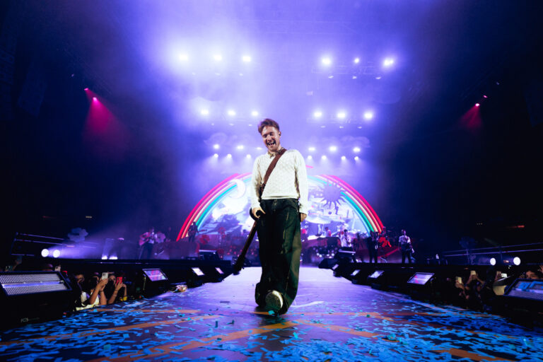il cantante Alfa su pedana davanti al palco durante un concerto; luci sul soffito e arcobaleno sullo sfondo
