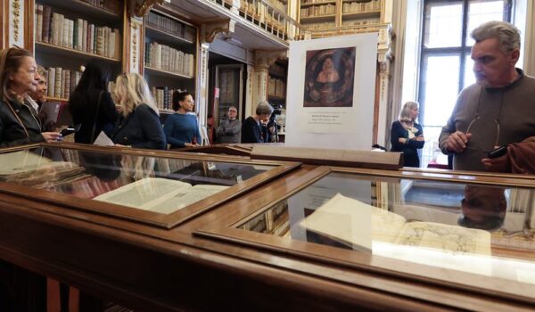 Biblioteca Moreniana durante una esposizione: teca con volumi antichi in primo piano, alle pareti scaffalature con volumi, pannello informativo e finestra a destra, visitatori