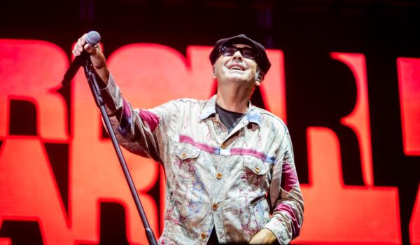 Luca Carboni durante un concerto a Genova, alle spalle una scritta rossa su sfondo nero, tiene in mano il microfono