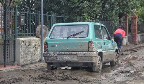 macchina e strada infangata in primo piano, sullo sfondo una donna intenta a spalare il fango