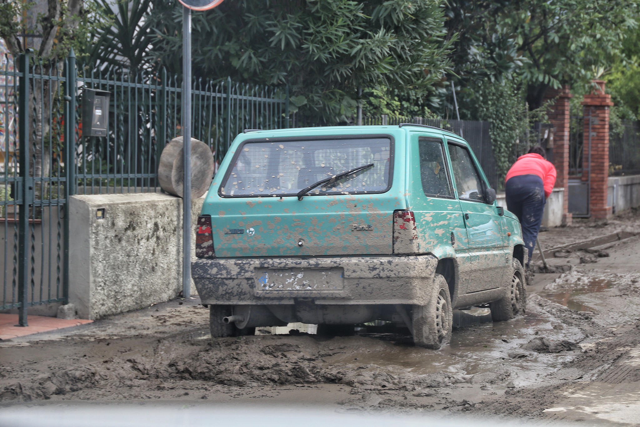 macchina e strada infangata in primo piano, sullo sfondo una donna intenta a spalare il fango