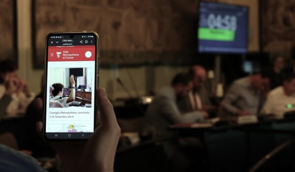 in primo piano cellulare sull'homepage del sito CMF, sullo sfondo la sala iv stagioni durante il consiglio metropolitano