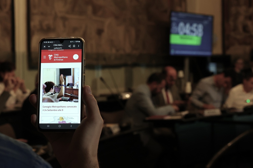in primo piano cellulare sull'homepage del sito CMF, sullo sfondo la sala iv stagioni durante il consiglio metropolitano